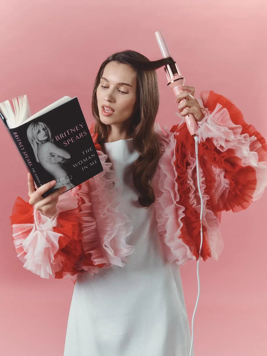 Woman holding a book and a hair curler against a pink background